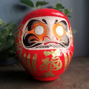 Front view of a traditional Japanese red Daruma doll featuring gold fortune lettering, white unpainted eyes and bold black and orange facial artwork.